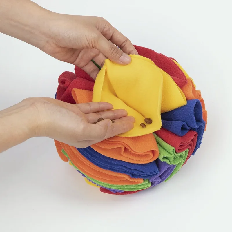 Hands lifting a yellow fleece flap on the rainbow ball to show kibble tucked underneath